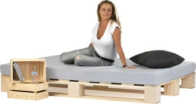 A woman sits on a bed frame made of wooden pallets, with a mattress and a pillow.