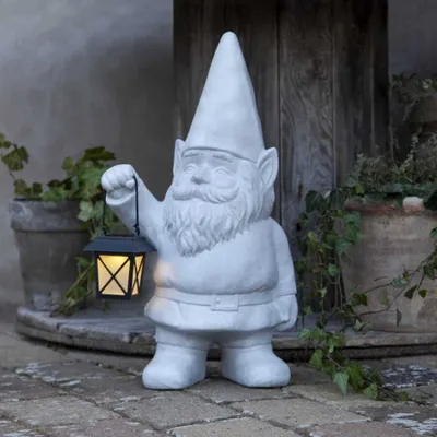 A garden gnome statue holding a solar lantern, placed outdoors on a stone patio with potted plants in the background.