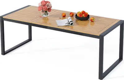 A rectangular outdoor dining table with a slatted wooden top and a dark metal frame, set against a white background. Decorative items like a vase of flowers, a bowl of fruit, and a magazine are placed on the table.