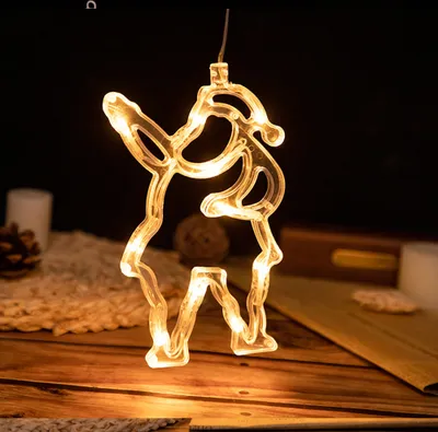 A close-up of a hanging LED Christmas decoration shaped like a reindeer, illuminated with warm yellow lights, set against a dark background with some blurred elements of a festive scene.