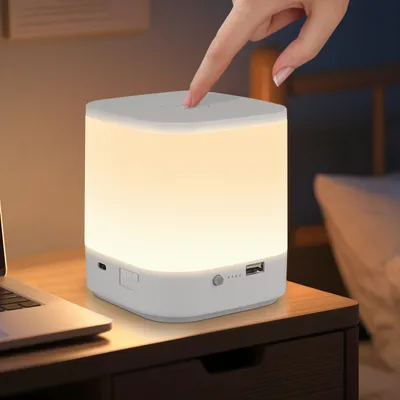 A hand is touching a white LED night light on a wooden nightstand next to a laptop.