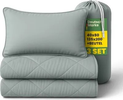A folded pillow and two folded blankets are stacked on top of each other, with a cylindrical bag standing next to them. The bag has a label with product information.