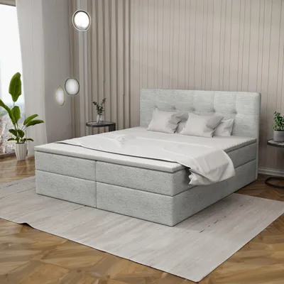 A bedroom scene featuring a large boxspring bed with a mattress and topper, set against a backdrop of textured walls and curtains, with a plant and side table visible.