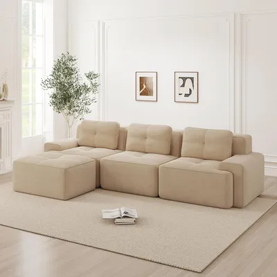 A beige modular L-shaped sofa with an ottoman is shown in a well-lit, modern living room setting.
