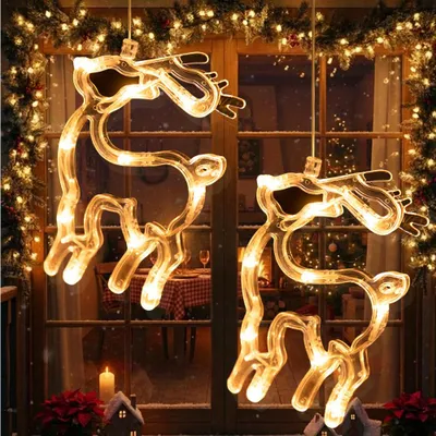 Two illuminated reindeer window decorations hang in front of a window, with Christmas lights and decorations visible in the background.