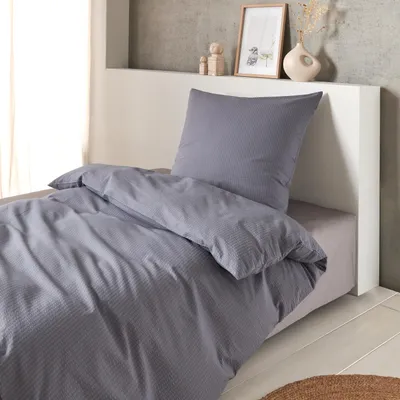 A bed with grey seersucker bedding and a pillow, set against a white headboard and a textured wall.