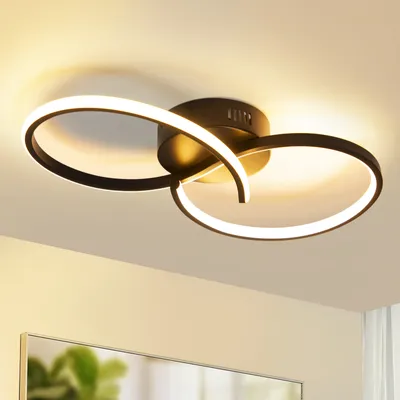 A modern black ceiling light with two intertwined rings, illuminated with warm white light, mounted on a white ceiling.