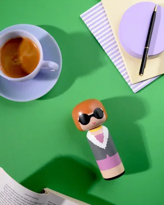 A stylized doll figure is placed on a green surface next to a cup of coffee and some stationery.