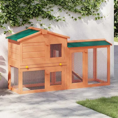 A wooden rabbit hutch with a green roof is placed on a paved patio next to a white wall with ivy growing on it.