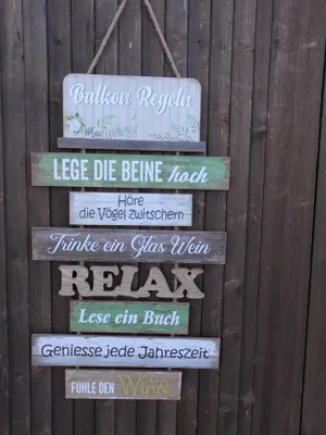 A decorative wooden sign with multiple planks, each displaying a rule or saying for a balcony, hanging on a wooden fence.