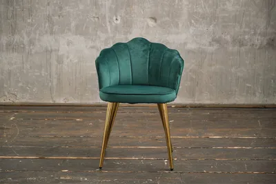 A green velvet chair with gold legs stands on a wooden floor in front of a concrete wall.