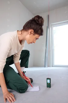 A woman is kneeling on a construction site, using a laser distance measurer and a pencil to mark measurements on a piece of paper.