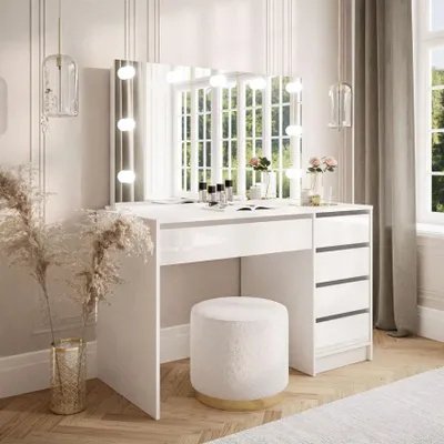 A white vanity table with a lighted mirror and drawers, a stool, and decorative items in a well-lit room.