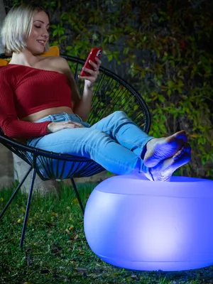 A woman is sitting in a chair outdoors at night, using her phone, with her feet resting on a glowing stool.