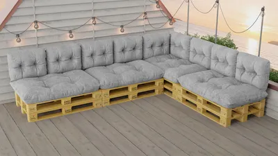 A sectional outdoor sofa made of wooden pallets with gray cushions, set on a wooden deck with string lights overhead.