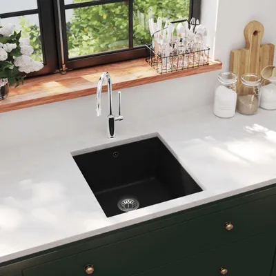 A modern kitchen sink with a black granite basin and a chrome faucet, set against a white countertop and dark green cabinets. A window with greenery is visible in the background.