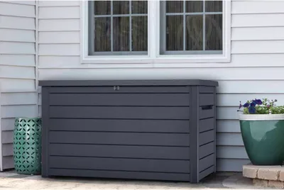 A dark gray storage box sits on a patio next to a house, with a green decorative stool and a potted plant nearby.
