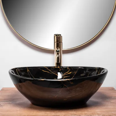 A black marble sink with gold veins sits on a wooden surface in front of a round mirror and a gold faucet.