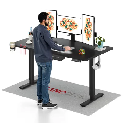 A man is standing at a black standing desk with three monitors, a keyboard, a mouse, and various desk accessories. A mat with a logo is on the floor in front of the desk.