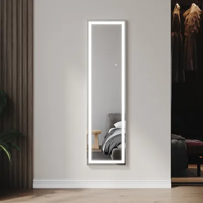A full-length LED mirror is mounted on a wall in a bedroom, reflecting a bed and a wooden stool. To the right, a closet with clothes is visible.
