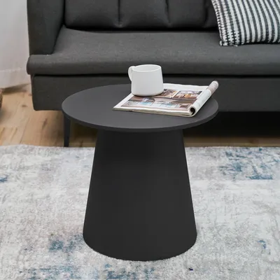 A round, black side table with a magazine and a mug on top, placed on a patterned rug in front of a gray sofa.