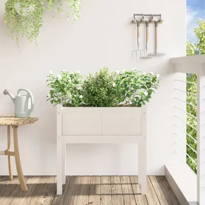 A white wooden planter box with plants sits on a wooden deck next to a small table with a watering can. Gardening tools hang on the wall behind it.