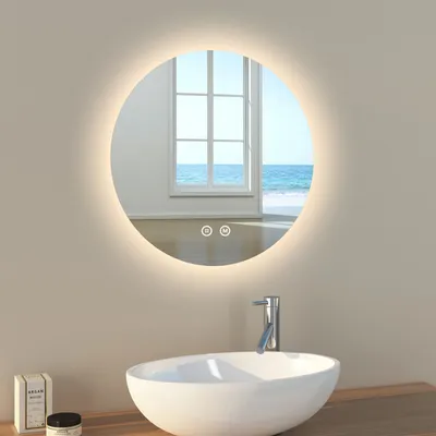 A round LED mirror with a light ring is mounted on a wall above a sink in a bathroom. The mirror reflects a window with a view of the ocean.
