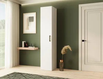 A tall white cabinet stands in a room with green walls, next to a white door. A small shelf with books and a framed picture hang on the wall above it. A vase with dried grass is placed next to the cabinet.