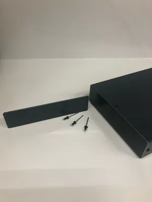 A dark gray metal end piece for a wall covering and three rivets are placed on a white surface next to a dark gray metal box.