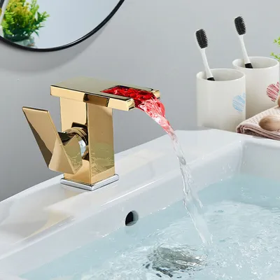 A gold waterfall faucet with red LED lights is shown running water into a white sink. Toothbrushes in cups are visible in the background.