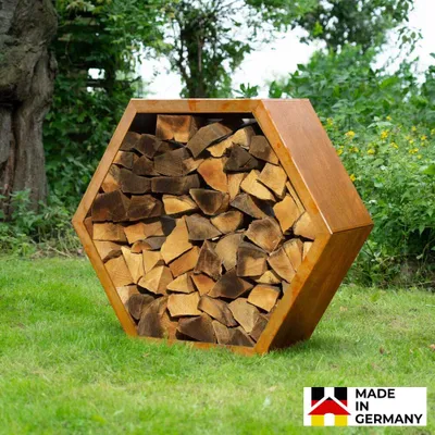 A hexagonal log holder filled with firewood sits on a grassy lawn, with trees and foliage in the background.