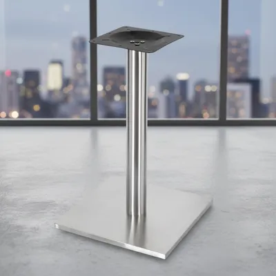 A table base with a square foot and a cylindrical column, placed on a concrete floor in front of a large window overlooking a city skyline at dusk.