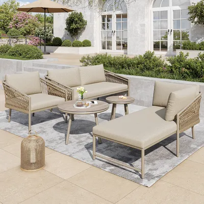 An outdoor patio scene featuring a five-piece garden lounge set, including a sofa, armchair, chaise lounge, and two coffee tables, set on a rug with a backdrop of a building and landscaping.