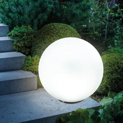 A glowing white sphere light sits on a stone pathway next to concrete steps and surrounded by lush green foliage and bushes in a garden setting at dusk.