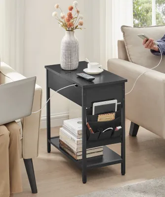A black side table with integrated charging ports and storage pockets is placed next to a sofa and an armchair. A laptop is on the armchair, and a person is using a phone connected to the table's charging port.