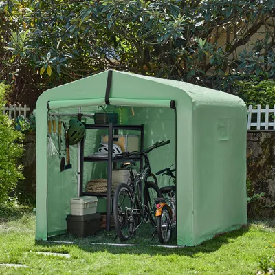 A green fabric garage for bicycles is set up in a grassy yard with trees and a white picket fence in the background. The garage contains two bicycles, shelves with storage bins, and gardening tools.