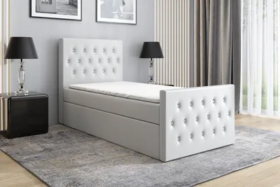A white boxspring bed with a tufted headboard and footboard, placed in a bedroom setting with bedside tables and lamps.
