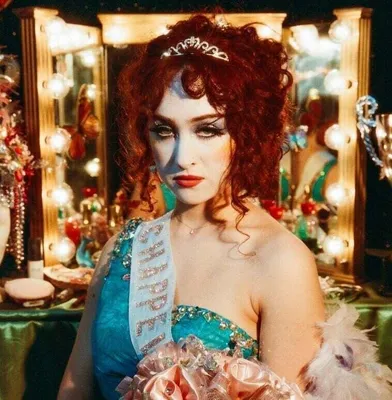 A woman in theatrical makeup and costume, wearing a tiara and a sash, poses in a dressing room setting with vanity lights.