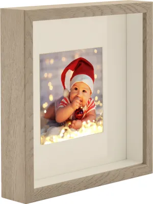 A baby wearing a Santa hat and a striped outfit is lying down and holding a red ornament in a shadow box frame.