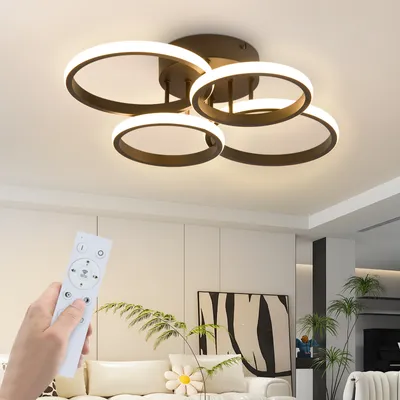 A hand holding a remote control in front of a modern ceiling lamp in a living room.