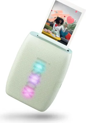A mint green Instax mini Link 3 photo printer is shown at an angle, printing a photo of a woman holding a heart-shaped balloon. The printer has three illuminated lights in blue, pink, and purple.