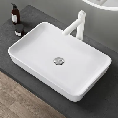 A white rectangular sink with a silver drain sits on a dark gray countertop. A white faucet is positioned behind the sink. To the left of the sink are two dark bottles, one with a pump dispenser.