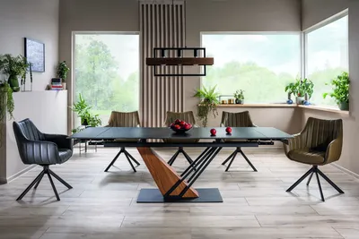 A modern dining room with a large extendable table and chairs, set near large windows with a view of greenery.