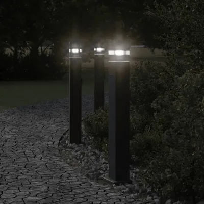 Three black bollard lights illuminate a cobblestone path at night, with bushes and trees in the background.