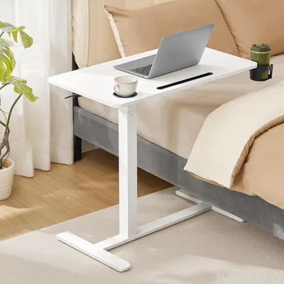 A white adjustable height table with a laptop, cup, and drink holder is placed next to a bed with tan bedding.