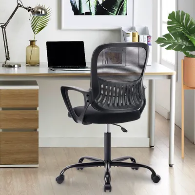 A black ergonomic office chair is shown in a home office setting, with a desk, laptop, and plant visible.