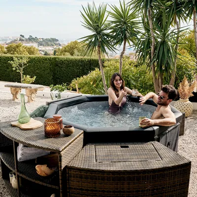 A couple relaxing in a hot tub outdoors, with furniture and plants around them.