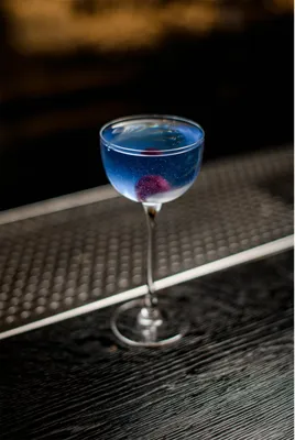 A blue cocktail with a cherry garnish in a coupe glass on a bar.