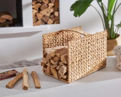 A woven basket filled with firewood, placed on a surface next to some matches and sticks, with a plant in the background.