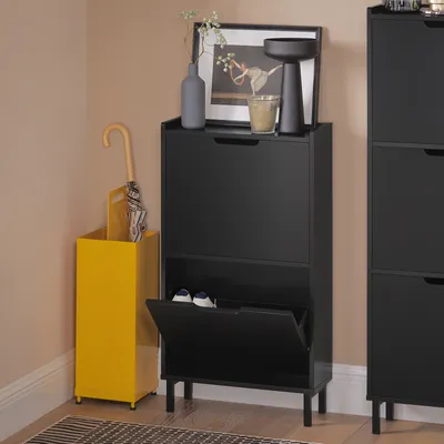 A black shoe cabinet with two flip-down drawers is shown next to a yellow umbrella stand. The top of the cabinet displays a framed picture, a vase, and a candle holder.